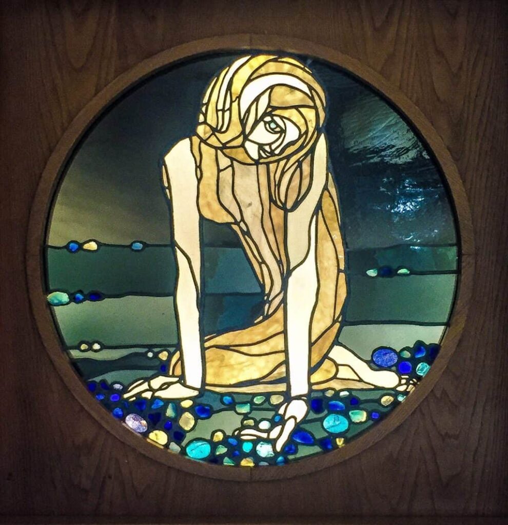 A stained glass window in the Owl House where a a woman with long blond hair is reaching down to the coean and picking up pebbles inside one of the coolest places in Rome. 