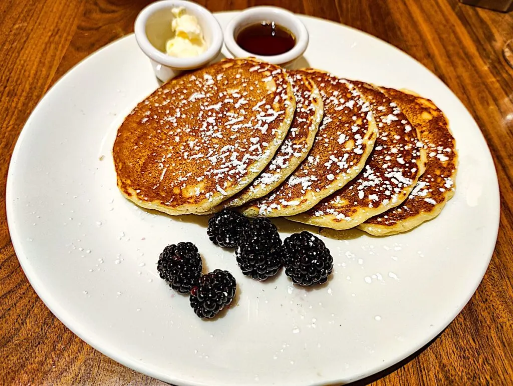 Five lemon ricotta pancakes sit on a white place with butter and syrup on the side as well as five blackberries. This is one of the best brunch places in NYC near the Freehand. 
