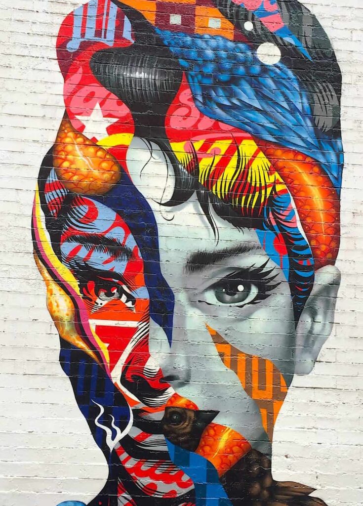 View of street art in Little Italy. It shows a black and white mural of Audrey Hepburn on a white wall that is bedazzled with vibrant colors during one of the best night tours in New York City. 