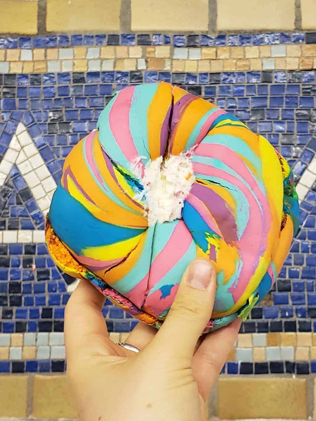 A rainbow colored bagel stuffed with cream cheese held up before I enjoyed one of the most delicious and yet unusual things to do in NYC.