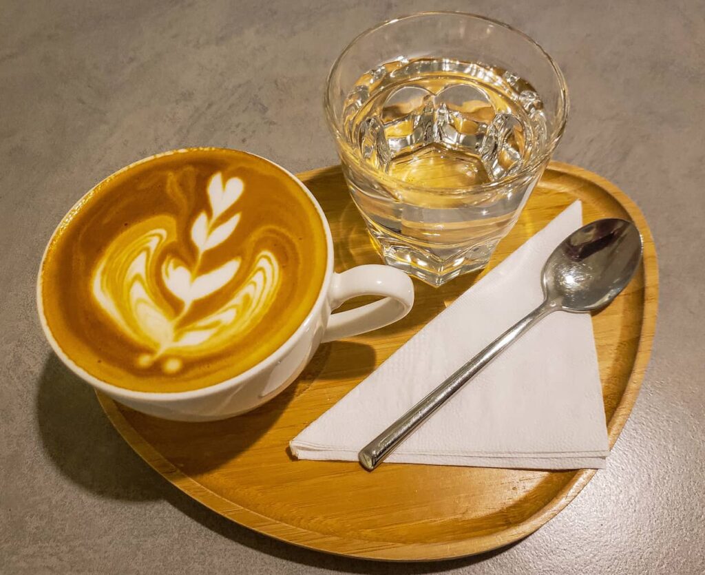S picture of a latte on a round tray with a glass of ice water on the side and a white napkin with a spoon.