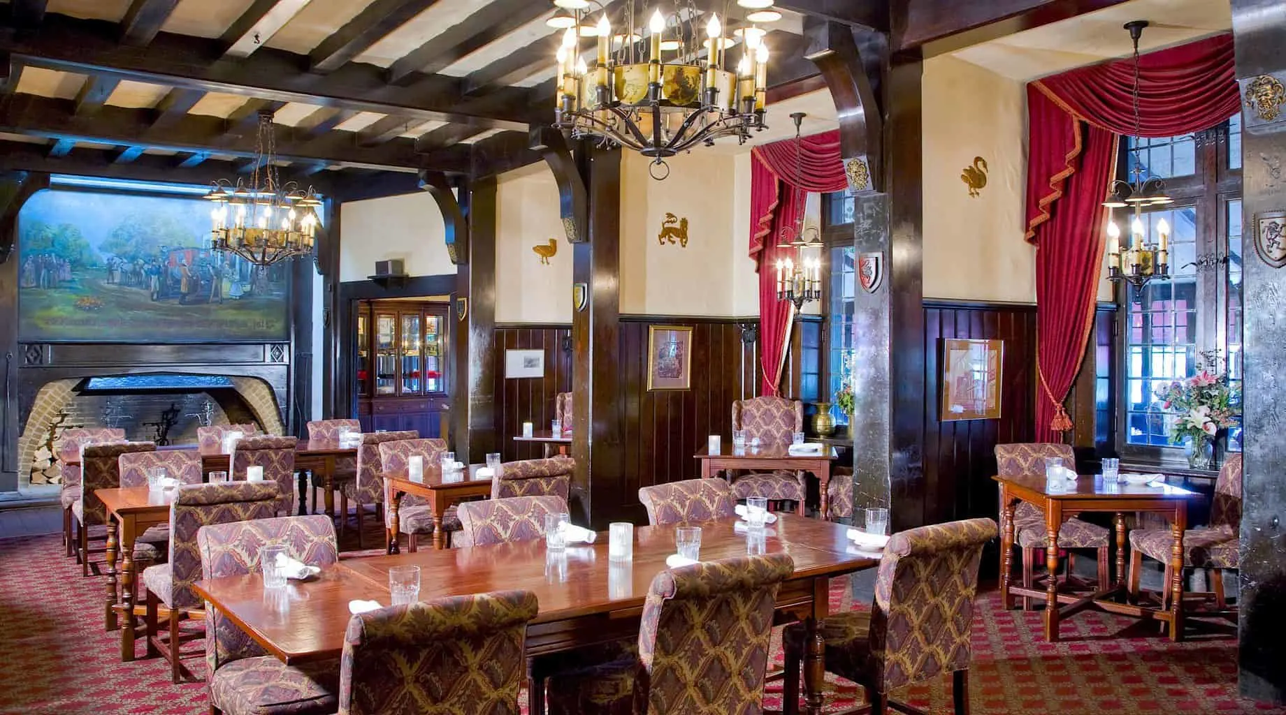 The refined and stately beamed ceilings, vibrant red curtains, upholstered chairs, and wood tables of the restuarant at the Red Coach Inn. 