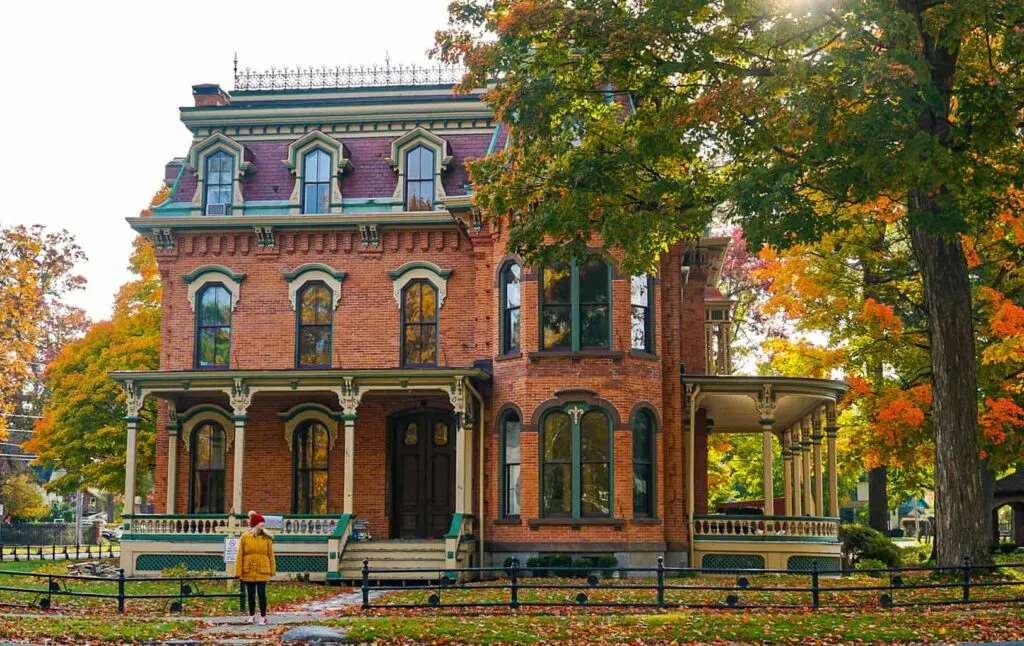 View of me stnding in front of a large, brick, Colonial-style home with vibrant foliage all around me in Saratoga Springs. 