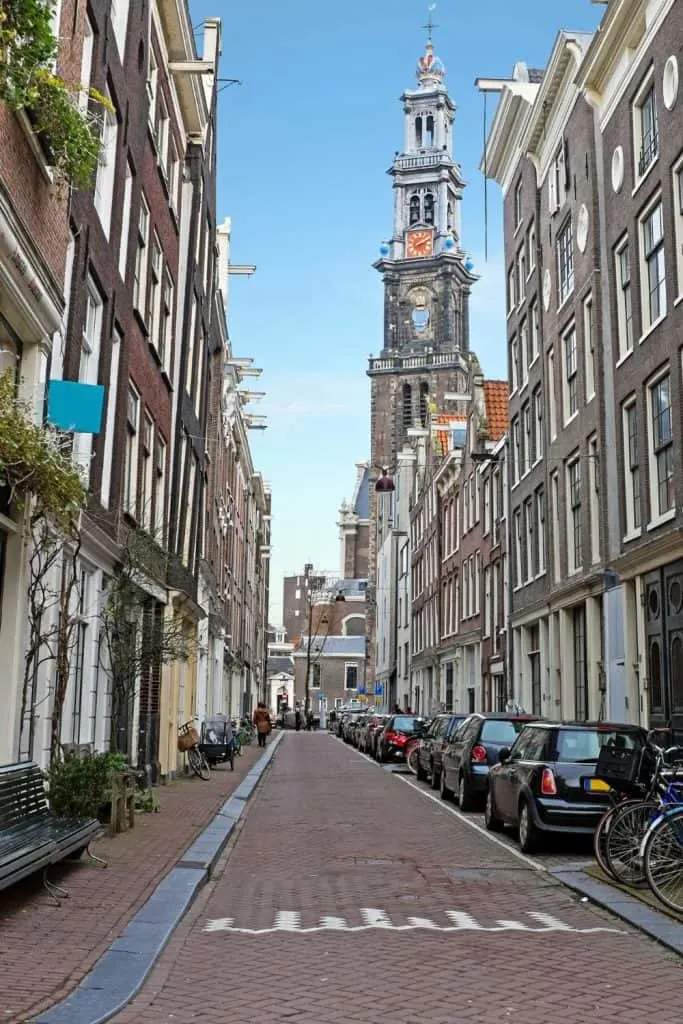 A view of the quaint homes that line the narrow but pretty streets of Jordaan. Cars line the streets and you see a church steeple in the background. 