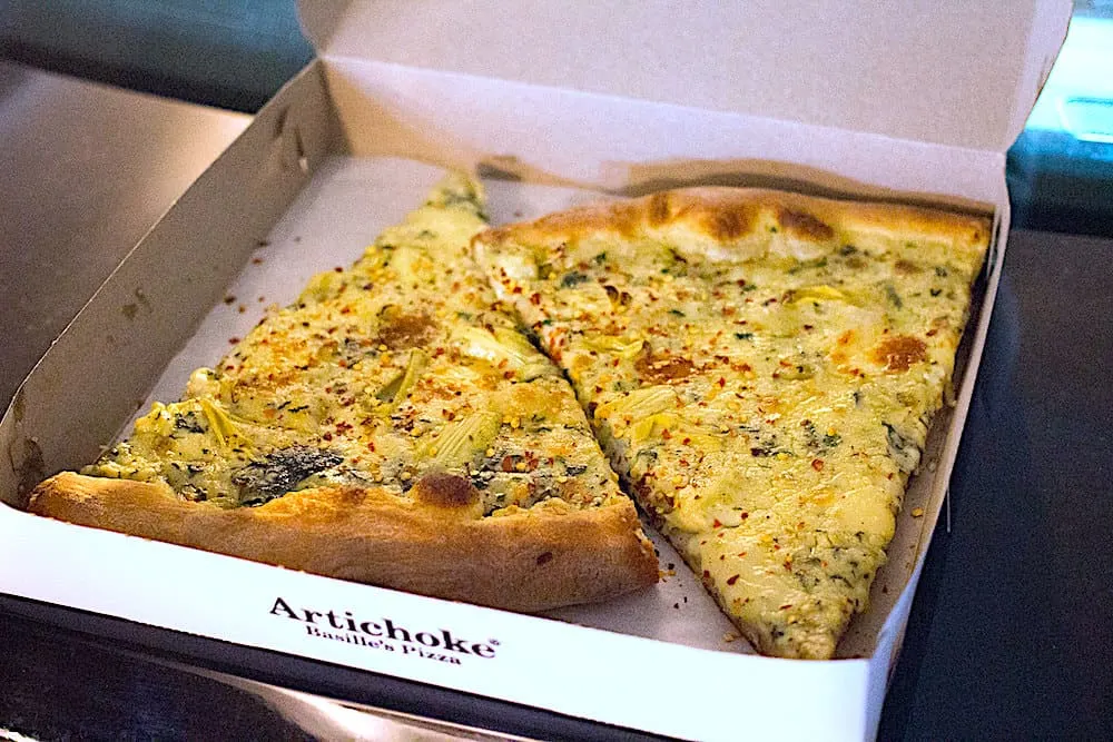 Two slices of artichoke spinach pizza in a pizza box from Artichoke Basille pizza.
