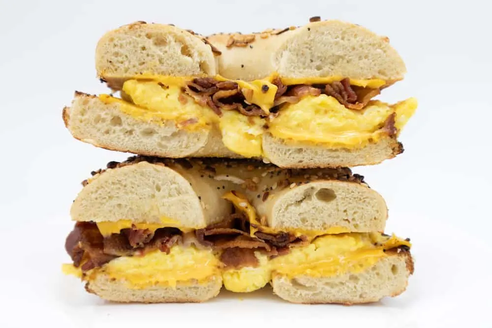 A cut in half side view of an everything bagel with bacon eggs and cheese on a white plate with a white background.