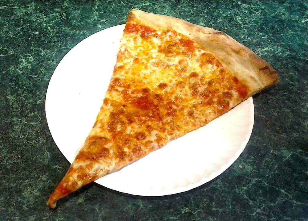 A classic slice of New York Pizza on a paper plate with a green table in the background.