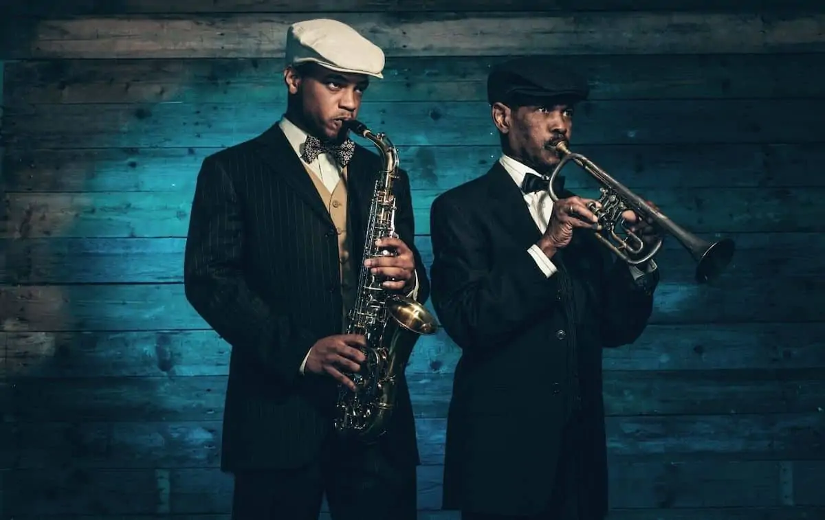 Two men playing jazz in a dark room with cabby hats and a sax and trumpet. 