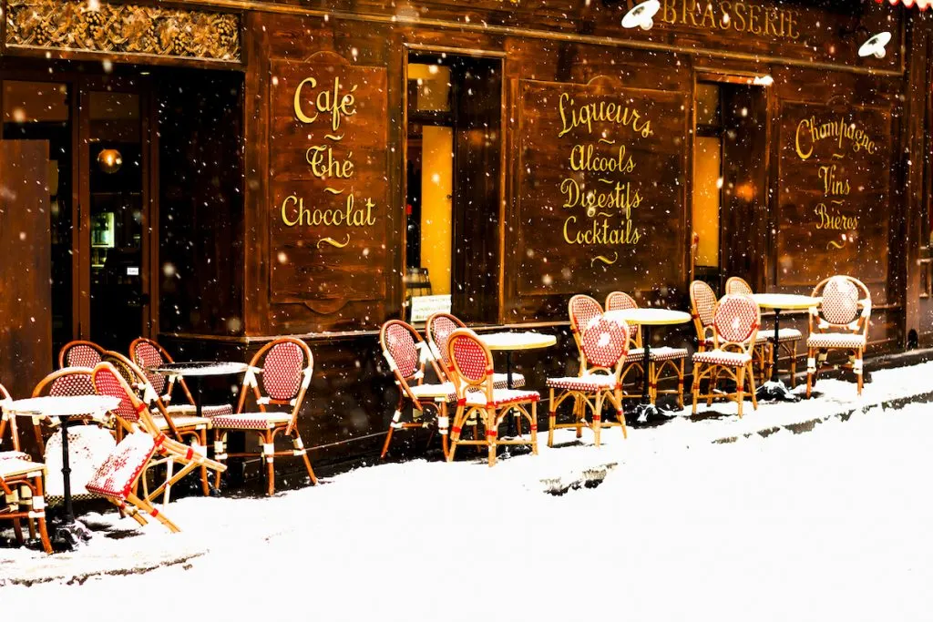 View of a Parisian cafe with red, whick seats outside and tables that are covered in snow with snow on the ground during winter in Paris. 