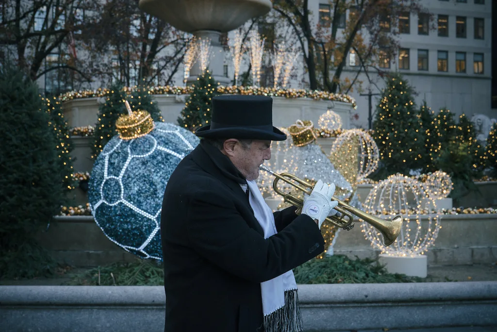 A street musician with a trumpet walking by the Pulitzer Fountain at Grand Army Plaza, which has some of the best Christmas lights in NYC. 