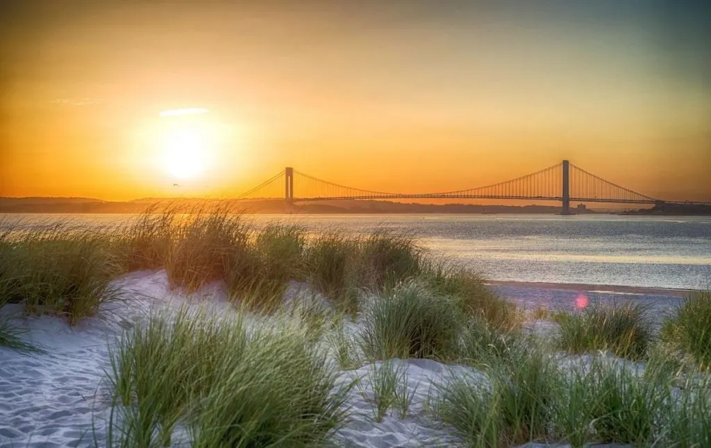 Sun setting over sand dunes in Brooklyn with the bridge in the background at one of the best beaches in New York City. 