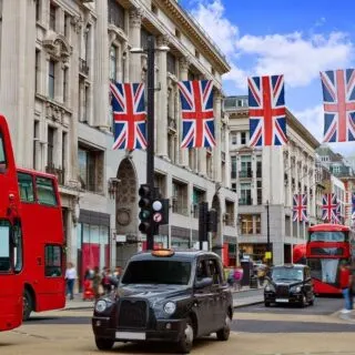 Double decker buses and black cabs traveling down busy Oxford Street with the flag hanging in the air along one of the most famous roads in London.