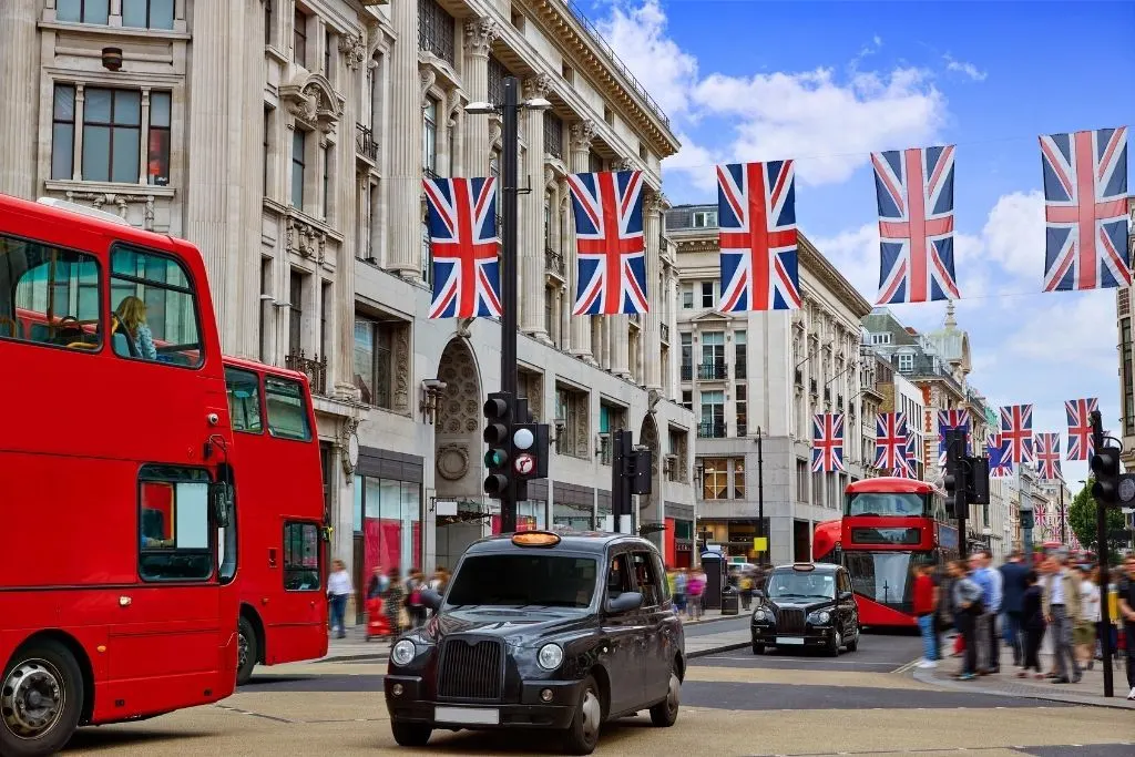 Double decker buses and black cabs traveling down busy Oxford Street with the flag hanging in the air along one of the most famous roads in London. 