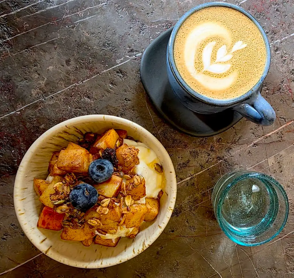 Aerial view of a cafe latte in a blaug mug and a small bowl of yogurt with fruit and granola on top. You can also see a water glass to the left. 