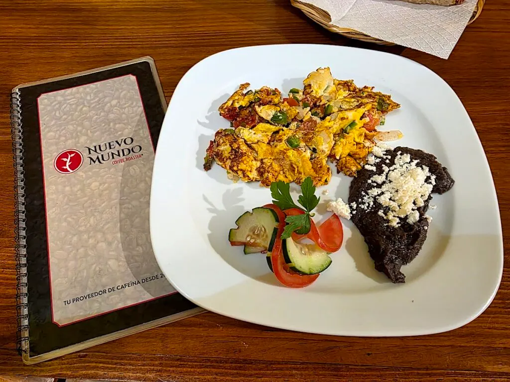 Aerial view of Mexican-style eggs with a small salad and refried beans on a white plate with the Nuevo Mundo menu to the left. 