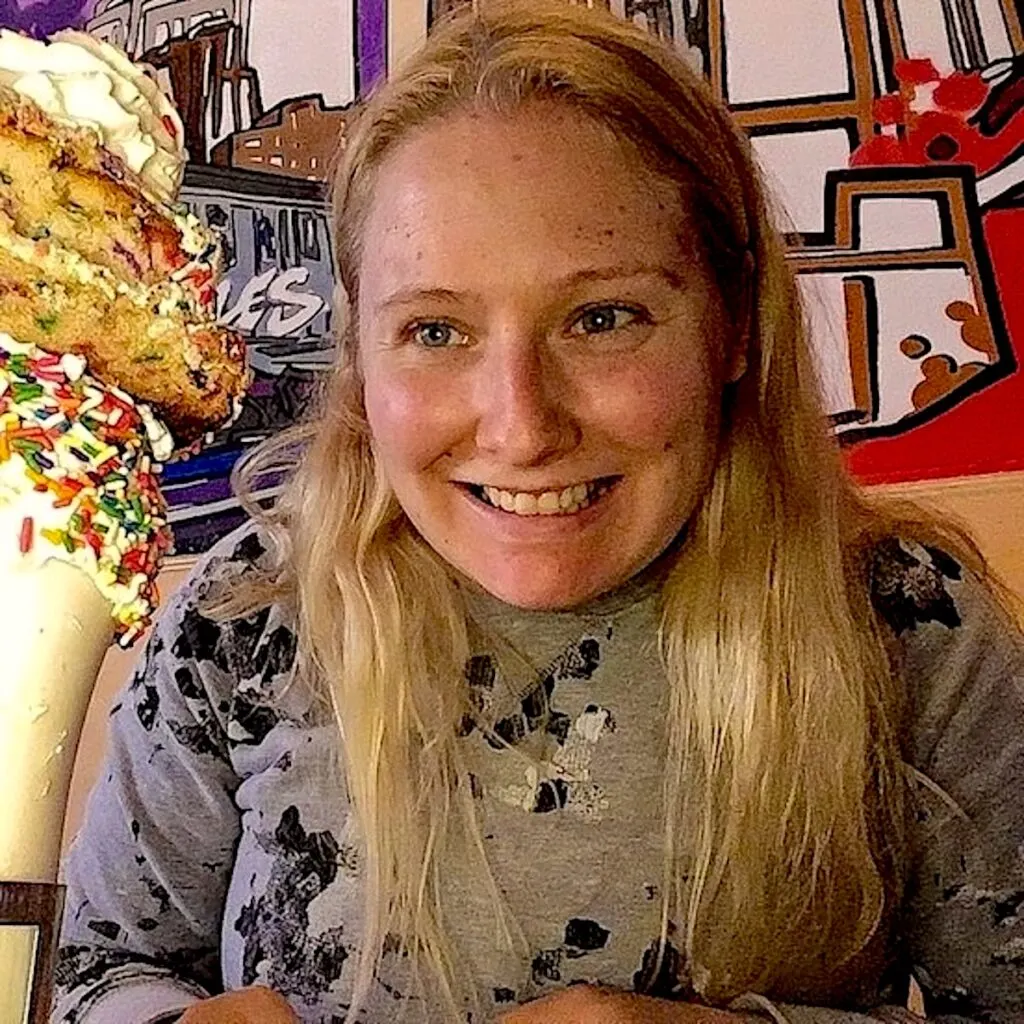 Me sitting in front of a giant milkshake with a piece of vanilla cake on top. I am sitting isnide Black Tap which has grafiti on the walls of one os the best dessert places in NYC. 