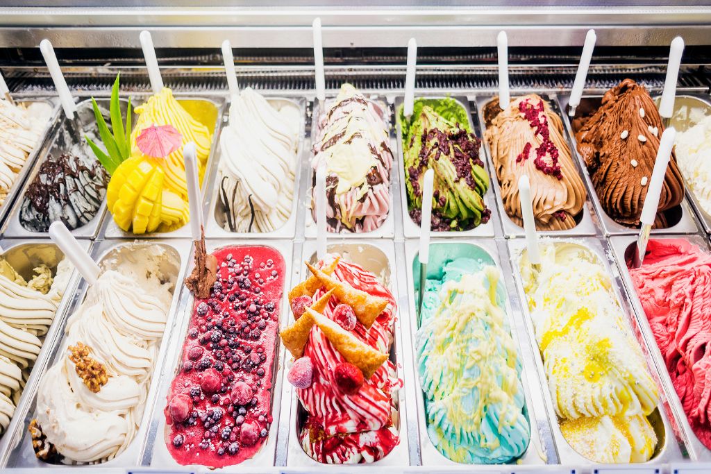 An assortment of different flavors of gelato in tubs with spoons sticking out. 