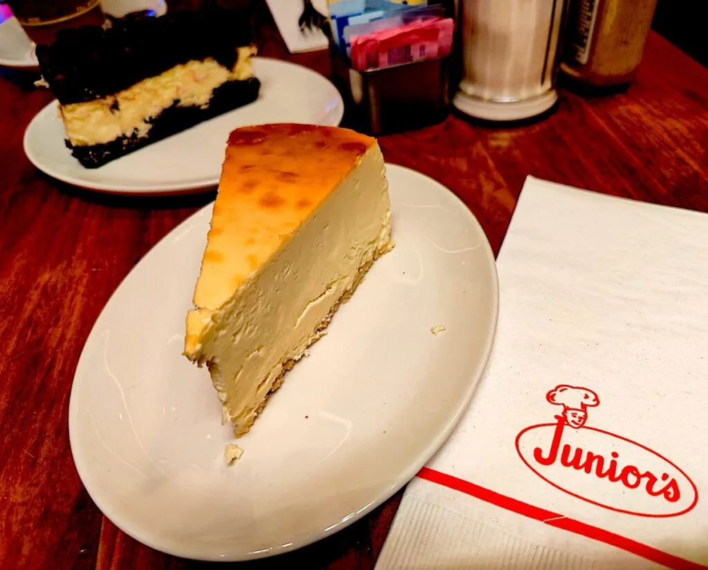 A view of a piece of original and brownie cheesecake on white plates. They sit on a wooden table and have a white napkin next to them at one of the best dessert places in New York City. 