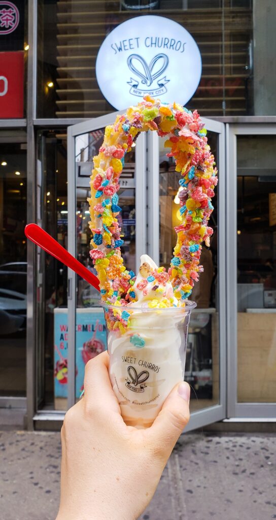 I am holding a cup of vanilla ice cream and a round churro covered in fruitty pebbles. There is a red spoon n the ice cream and I am standing in front of the Sweet Churros sign. 