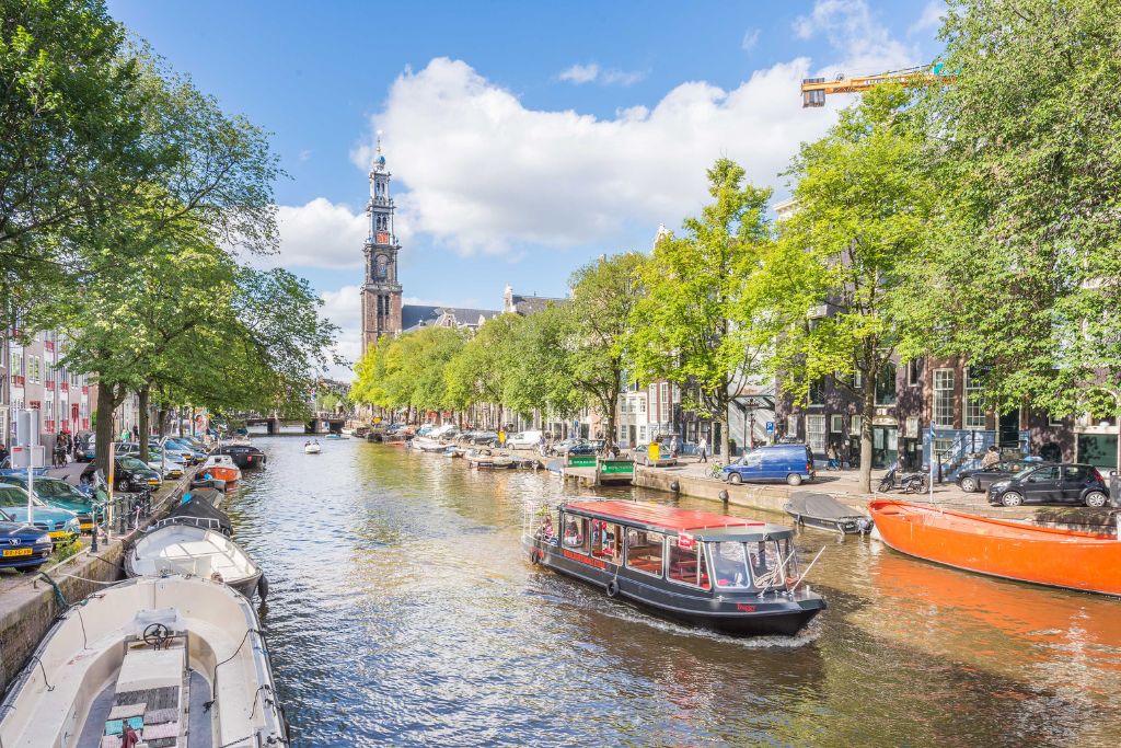 A view of canal boats running up and down Amsterdam. The trees lining the canals are green, the sky is blue, and there is a church steeple in the background. You can see all this during one of the bets food tours in Amsterdam. 