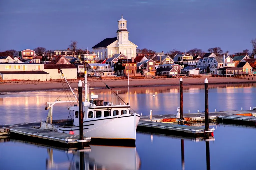 A fishing boat in the water, docked at a pier and a white church in the background in Cape Cod. 