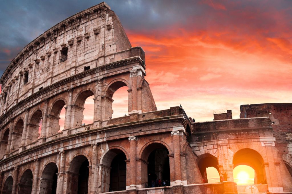 A stunning up close view of the Colosseum with the sun shining through the windows and setting in the background which is a pink/orange sky.
