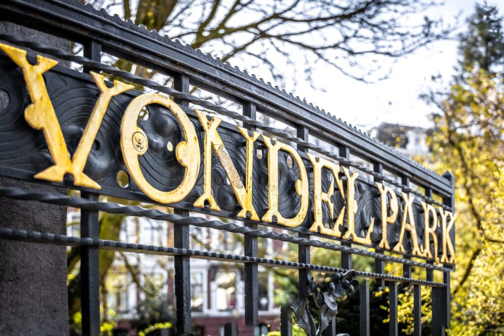 The black gate to Vondelpark with letters  in yellow that read "Vondelpark". There are also trees and houses in the background. 