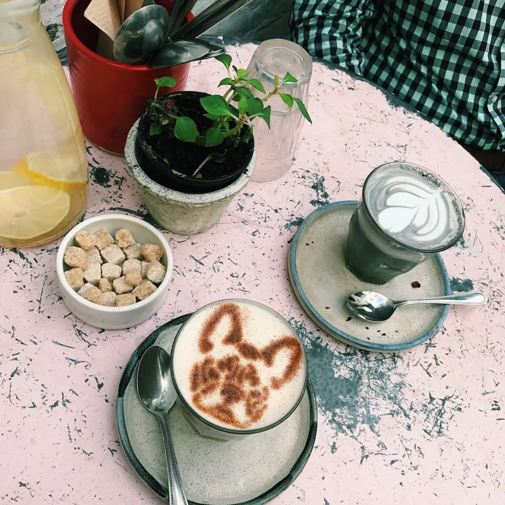 Aerial view of a latte with a dog shape on it and a charcoal latte sitting on a pink table at Farm Girl London. 