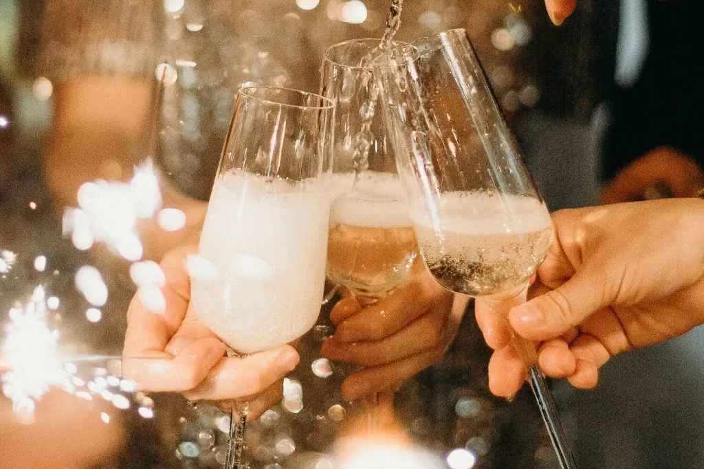 three people holding champagne flutes about half filled with champagne. They are holding their glasses together as champagne pours from above in the flute and sparklers glow in the background. 