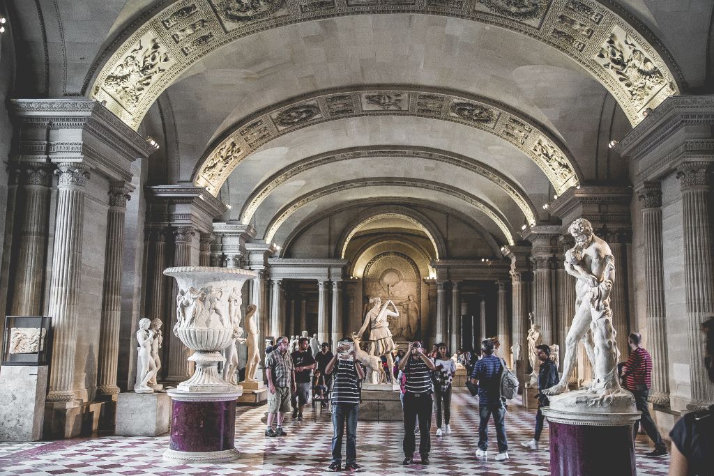 View of a gallery inside the louvre with classic white statues and tourists taking photos everywhere. 