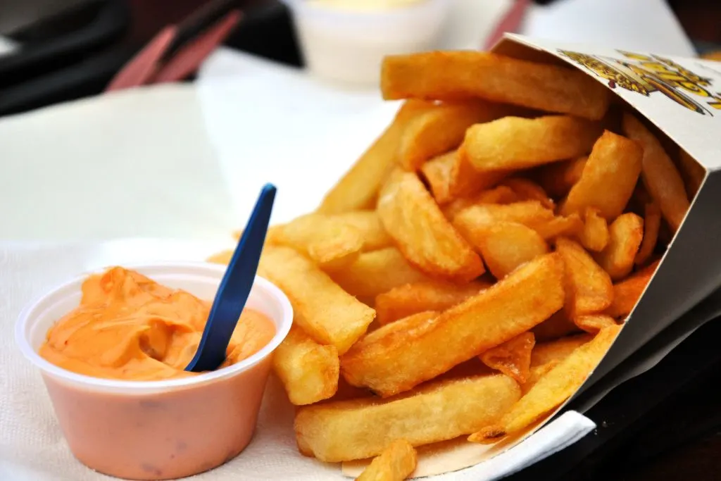 Thr best Belgian fries in Brussels spilling out from a take away container with pink dipping sauce on the side in a plastic cup that has a small spoon. 