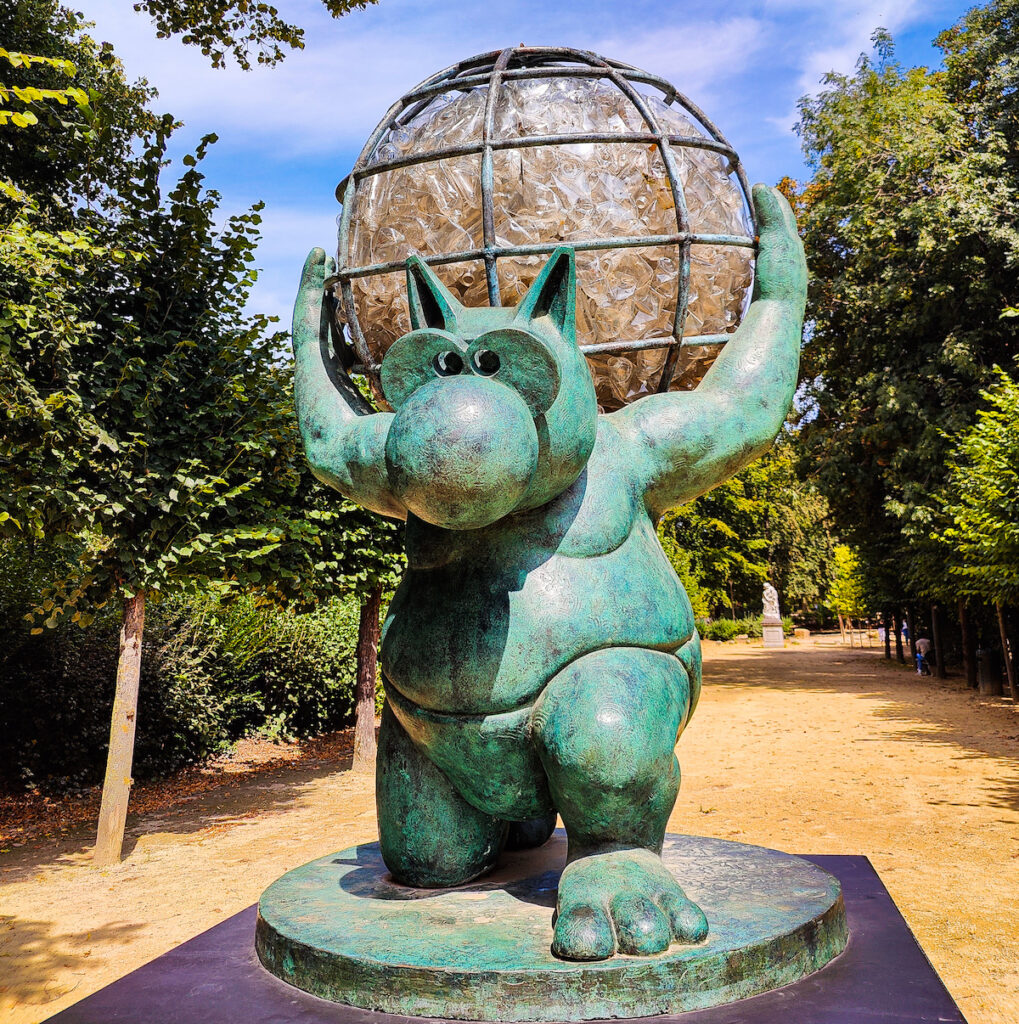 A view of a green-hued cat statue holding the world on its back in Brussels park like the Atlas Statue in NYC. The animal has one knee bent with dirt paths and trees around it.