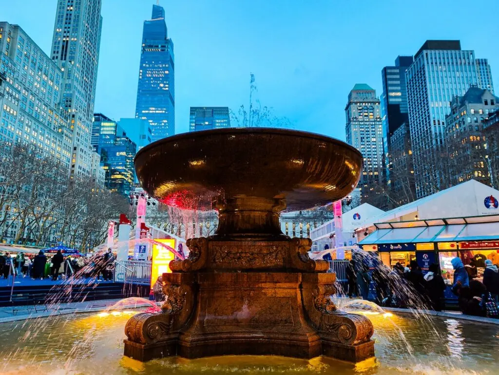 The fountain at Bryant Park in the evening with water coming out. It is surrounded by skyscrapers and there are buildings associated with the winter market at Bryant Park arund the fountain. 