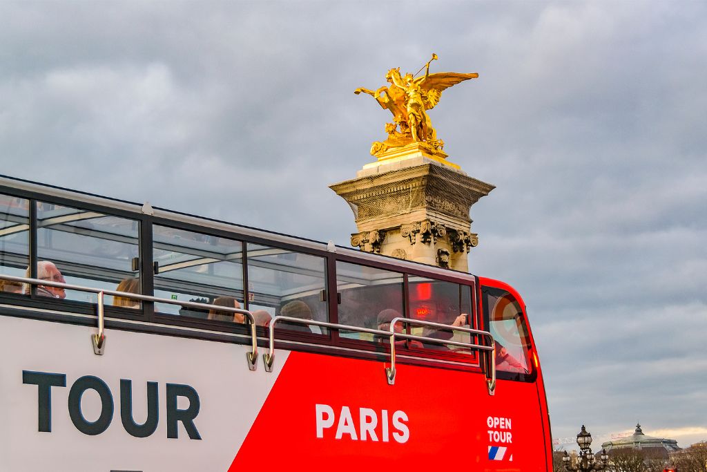 Red bus riding through the streets of Paris with a gold statue in the background and a gray sky. 