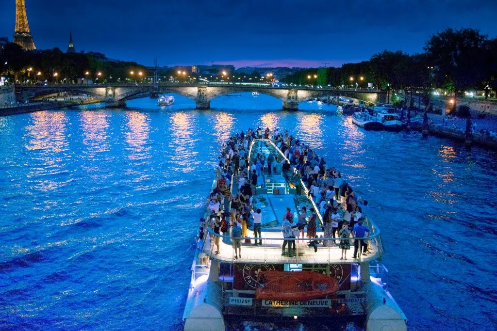 A cruise ship motoring along the Seine in the evening with the Eiffel Tower just visible on the right during one of the best night tours Paris has to offer.