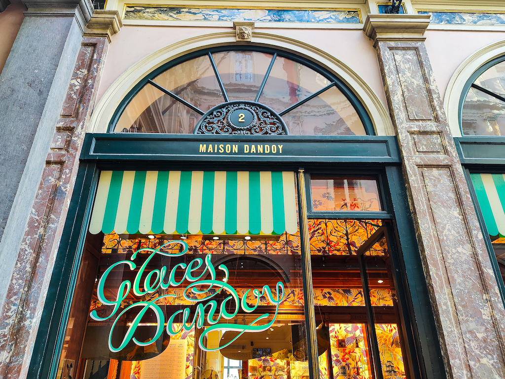 The exterior of Maison Dandoy in the Royal Gallery of Saint Hubert in Brussels. The exterior is surrounded by marbel and has a windo with green cursive writting that says Galces Dandoy since this is Maison Dandoy makes the best waffles in Brussels.