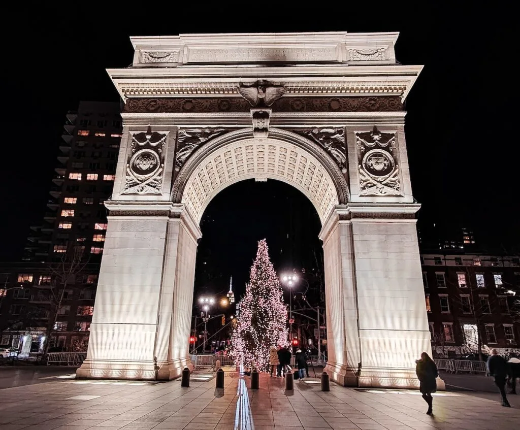 Stunning marble arch in Washington Square Park is all lit up in the evening. People walk pasr and there is a Christmas tree with white lights beneath the arch. 