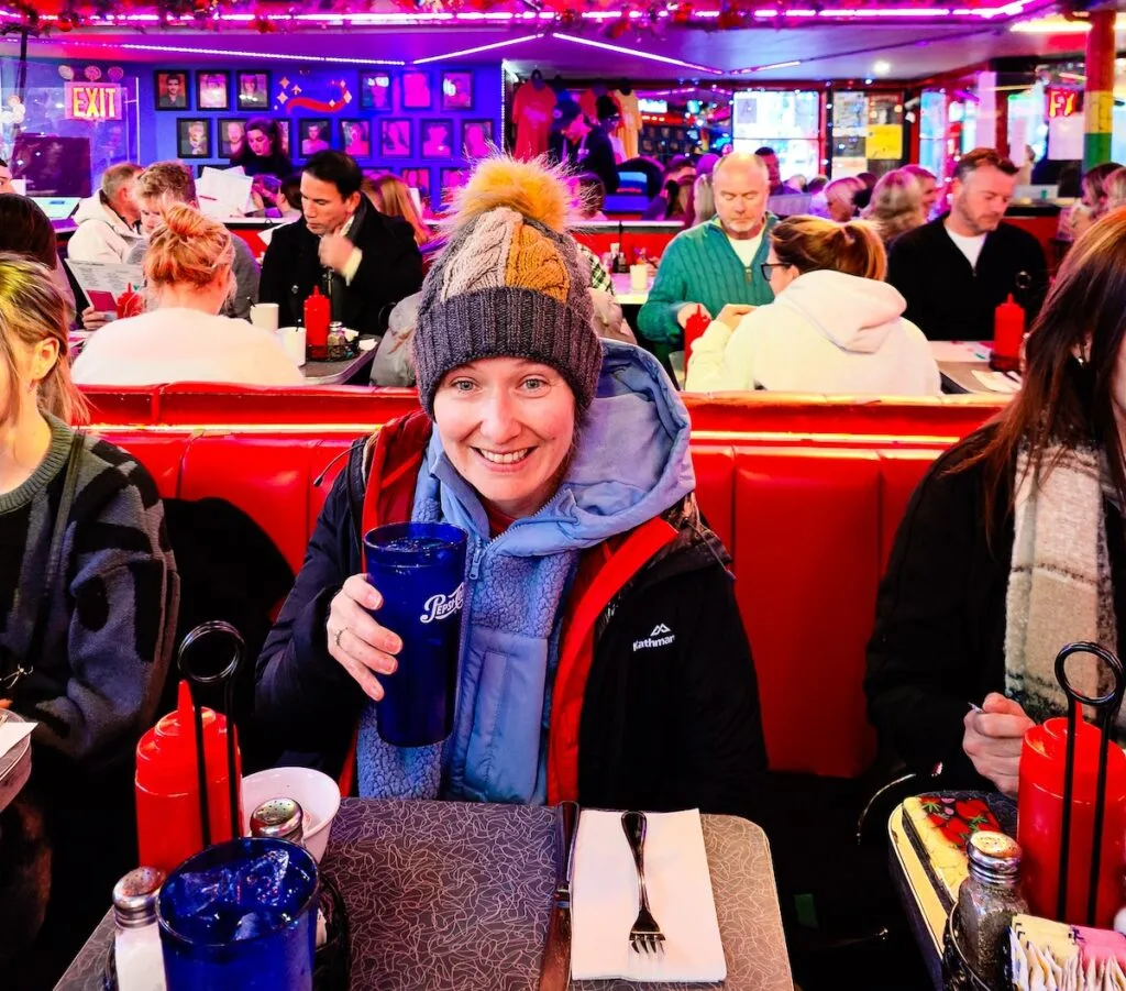 Me holding a blue glass of soda and wearing a black jacket, blue fleece, and winter hat. I am sitting in a red booth at one of the best themed restaurants in NYC.