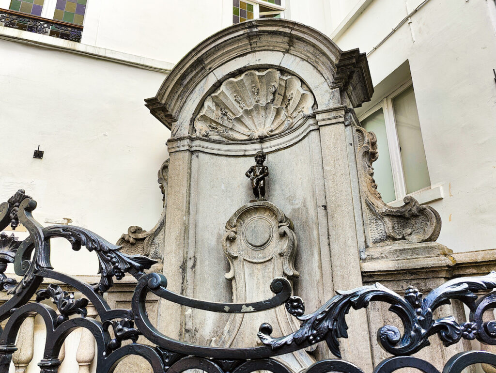 A view of Manneken Pis in the center of Brussels during your one day in Brussels itinerary. The statue of the peeing biy is behind a black fence and sits in a marble fountain. 