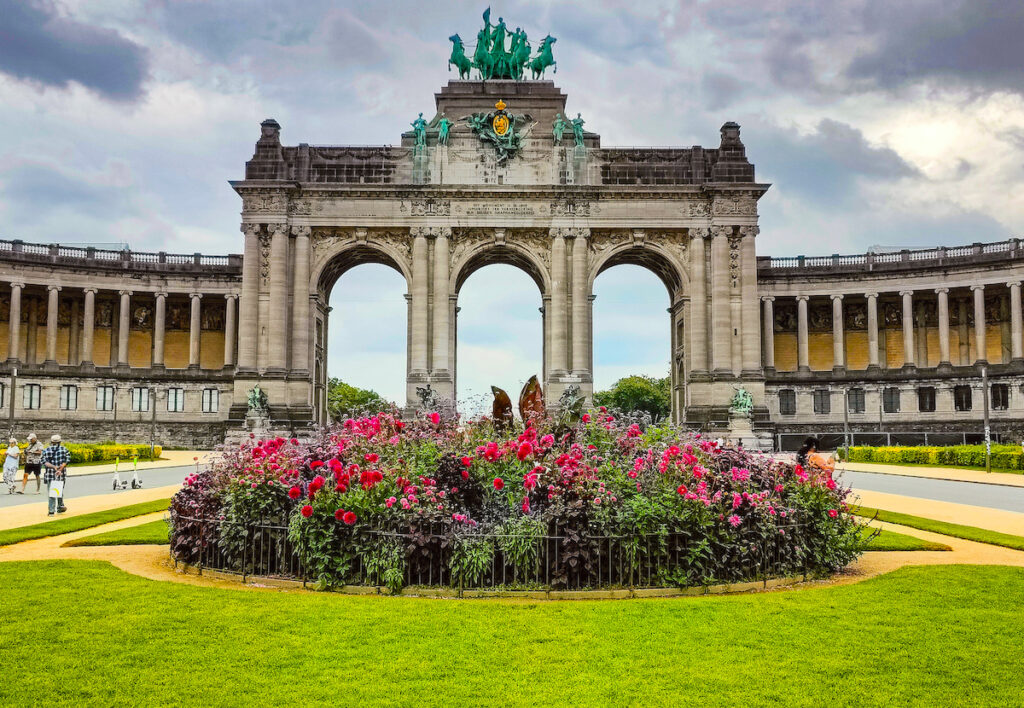 A view of the magnificent entrance arch at Parc du Cinquantenaire with 3 arches and horses on top. Flowers sit in front and there are clouds in the sky in the background.