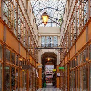 View of Passage du Grand Cerf. This is one of the best arcades in Paris with a glass roof, vintage light fixtures on the roof, and corridors lined with wood framed shops with glass store fronts.