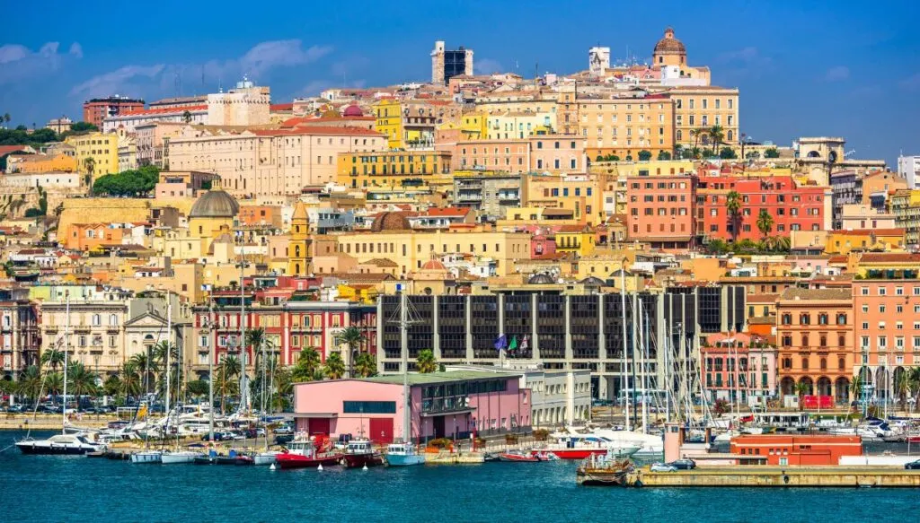 The vibrant homes that line the coast of Sardinia is the busy city of Cagliari. Boats also sit in the foreground and bob around in the blue water. 