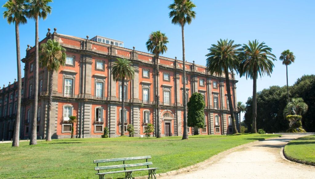 The stately pink exterior of Capodimonte Palace and Museum. It is surrounded by a luch park with green benches and walkways and has lots of tall palm trees. 