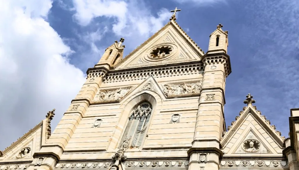 The white, stone exterior of the Cathedral of naples. Two small towers sit on either side of a central triangular rood with a circular window. The church features Gothic, Baroque, and Renaissance-style architecture and is a must-see during your Naples 1 day itinerary. 