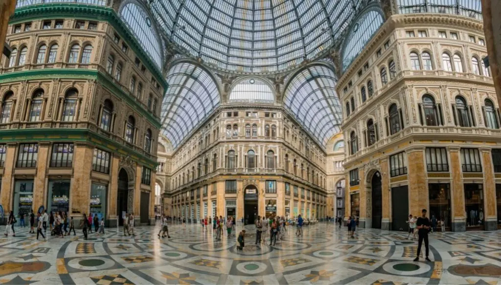 The impressive interior of Galleria Umberto I in Naples. It is a stunning, glass roofed shopping arcade with tiled floors and old world style buildings that house local shops. You can see people walking through one of five different pedestrian walkways here. 