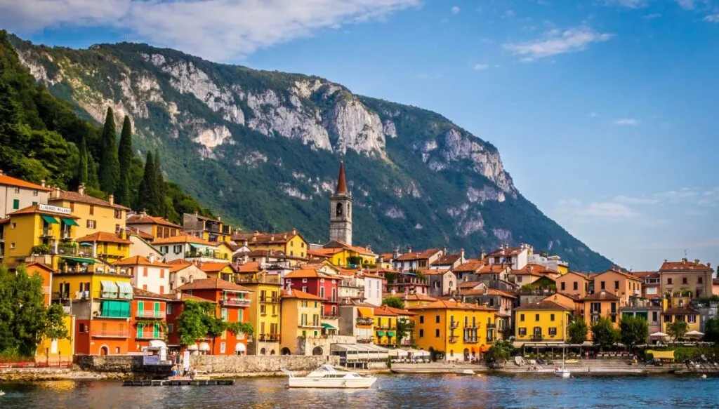 View of the vibrant orange and yellow homes and churches that line the coast of Lake Como. Mountains sit in the background and you can see all this during one of the best tours to Lake Como from Milan.