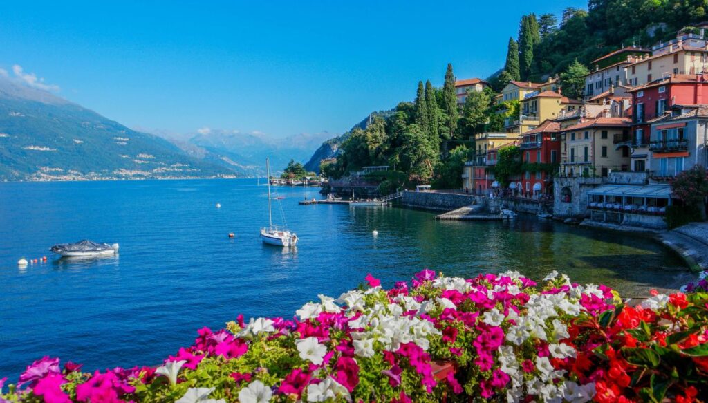 A view of the beautiful blue waters of Lake Como with mountains surrounding the lake with boats bobbing in the water. Colorful houses sit on the right shore and flowers line the bottom of the photo. This is what you'll see during one of the best day tours from Milan to Lake Como.