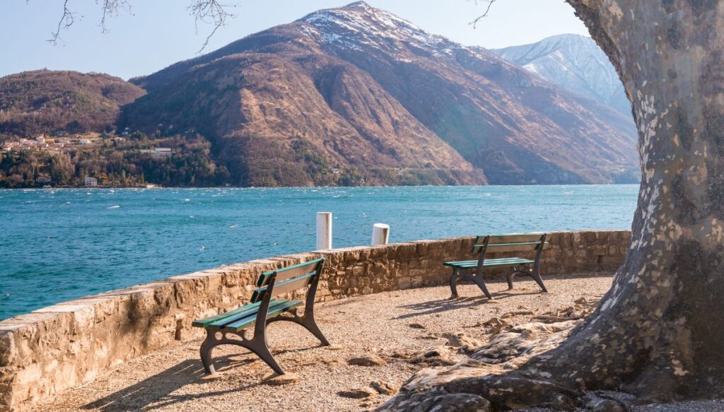 View of mountains covered in snow and the blue water of Lake Como beneath them by a lakeside promenade with two empty benches.