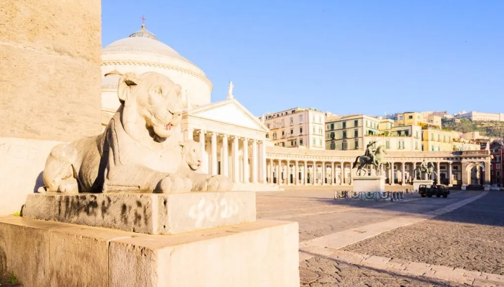 A view of Piazza del Plebiscito. You can see a white lion statue on the left and a giant Roman style church with a central dome and en entrance with columns and a trianular roof. The piazza is surrounded by a white semi-circle of columns and there is a dark statue of a man riding a horse on the right. A great place to visit during your Naples 1 day itinerary. 