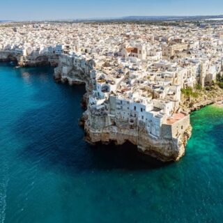 Aerial view of the town of Polignano A Mere in Puglia. It is a must-see when renting a car in Puglia. From this photo you can see white homes perched on a steep cliff that is surrounded by a beach and crystal blue water.